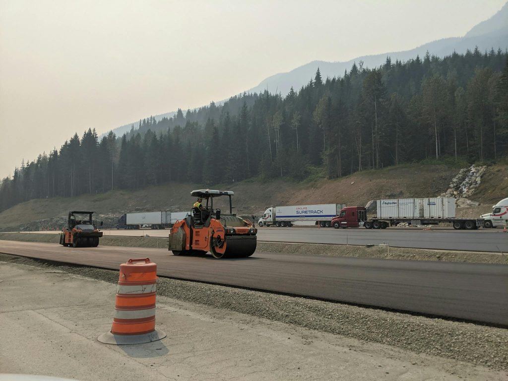 Asphalt road construction with heavy machinery and trucks in a scenic mountainous area.