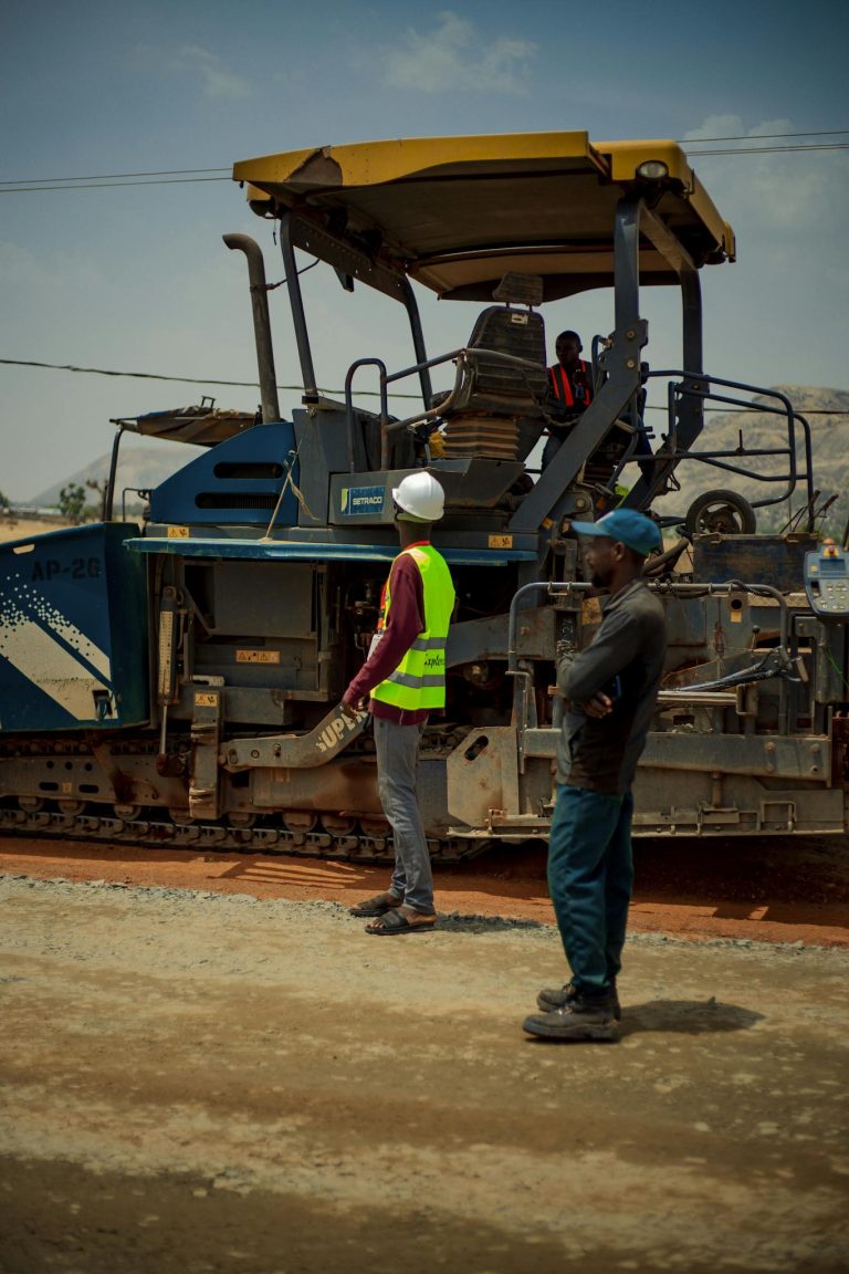 Two construction workers observing roadwork machinery under bright daylight.